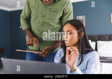 Diverse couple using laptop and tablet talking in bedroom Stock Photo