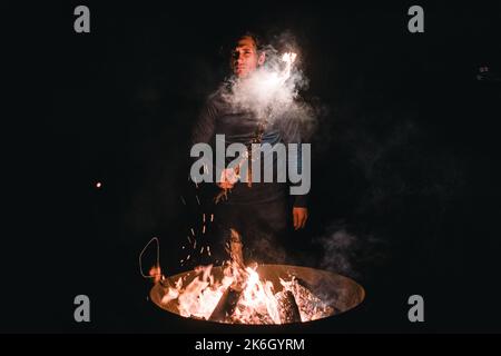 bonfire burning inside a large metal container and caucasian boy standing in a black t-shirt and dark pants seriously looking at camera and holding a Stock Photo