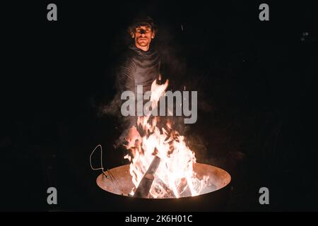 standing caucasian boy in a black t-shirt serious looking at camera stoking the fire inside a big metal pot full of firewood burning at night and very Stock Photo