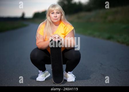 Chubby middle aged European woman, overweight, in a tracksuit with a skateboard, tired after playing sports, resting. Stock Photo