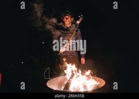 standing caucasian boy in a black t-shirt and dark pants seriously looking at camera with a dry branch of a burning tree near a large metal pot full Stock Photo