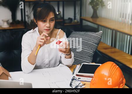 Beautiful young Asian woman holding house model while sitting in front of her working place. Stock Photo