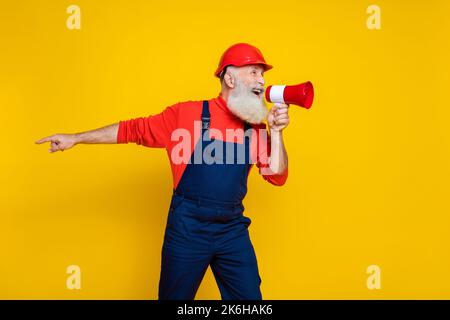 Photo of angry serious old guy dressed uniform overall red hardhat screaming toa pointing finger empty space isolated yellow color background Stock Photo