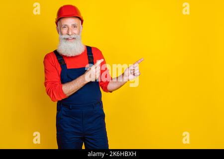 Photo of funny cheerful age man workwear overall red hard hat pointing fingers empty space isolated yellow color background Stock Photo