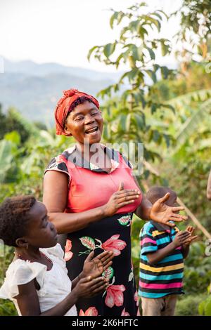 A happy African woman sings with her family outside her home in Uganda's Rwenzori Mountains, Kasese District, East Africa. Stock Photo