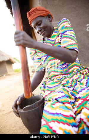 Elderly woman in traditional colorful dress pounding peanuts with a wooden mortar and pestle in food preparation in Uganda, East Africa. Stock Photo