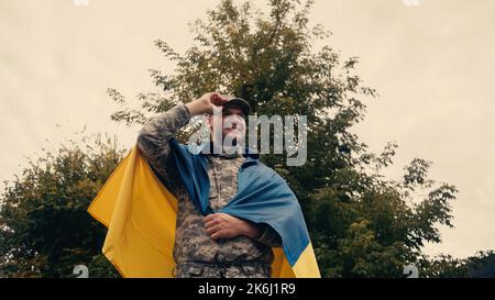 low angle view of happy soldier in military uniform adjusting cap while holding ukrainian flag outdoors,stock image Stock Photo