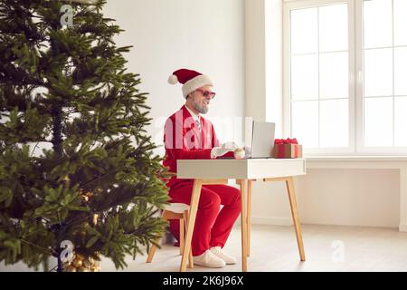 Funny man in Santa costume working on laptop computer in office with Christmas tree Stock Photo