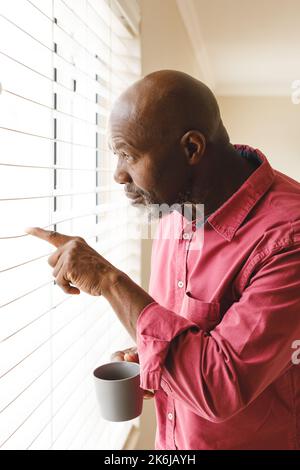 Vertical image of curious african american senior man looking out of window drinking coffee at home Stock Photo