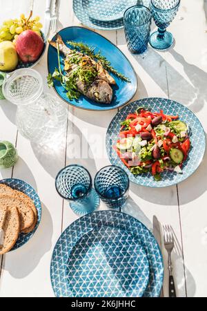 Gilt-head bream also known as Orata baked in the oven and served on a white wooden table with vegetable salad and fruits. Healthy seafood preparation Stock Photo