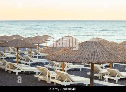 White color sunlounger chairs on black sand beach with natural reed sun umbrellas at sunset. Holiday in Greece concept, lot of copy space. Stock Photo