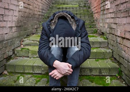 A lonely man in crisis, sitting on the steps of the old street. The concept of drug addiction and alcholism. Stock Photo