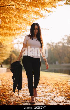 Young woman walking in park under trees with orange leaves during sunny weather in autumn Stock Photo