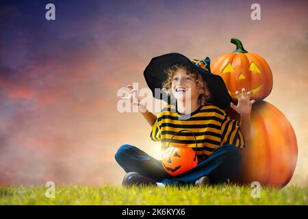 Little boy in witch costume sitting at giant pumpkin lantern on Halloween night. Kids trick or treat. Children have fun. Spooky and scary celebration. Stock Photo