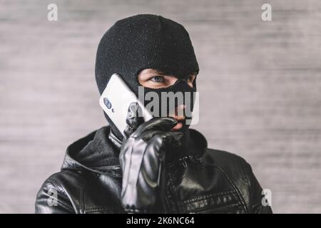 The thief is talking on the phone in a black mask. The extortionist demands money for the kidnapping. a phone fraudster commits a crime, violates the Stock Photo