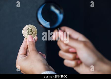 Close up of female hand holding magnifying glass and looking through it at bitcoin. Stock Photo