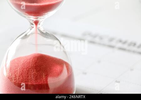 Close up of red sand slipping away through hourglass, time running out, December calendar out of focus in background, end of year, no more time Stock Photo