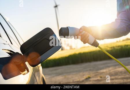 woman hand inserts power cable supply to charge electric or EV car at sunlight Stock Photo