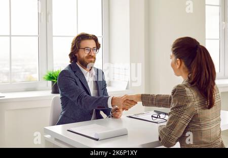 Happy business agent and client sitting at office desk, making deal, smiling and shaking hands Stock Photo