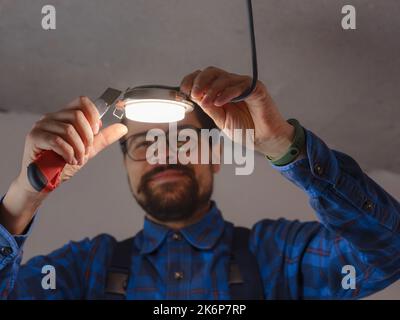 Electrician at work. Service for repair of electrical wiring and replacement of ceiling lamps. builder is installing small lamp on ceiling. Rent-a-gent helps with housework Stock Photo