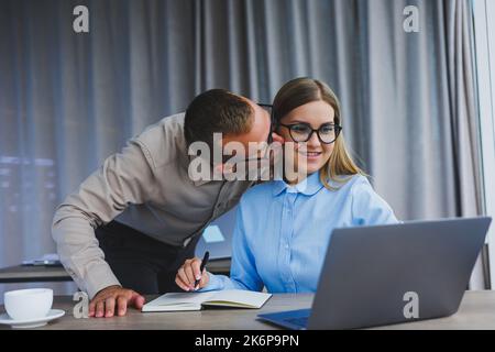 Young cheerful multinational corporate lawyers in formal wear editing text of new contract using laptop while sitting at table with notepad in modern Stock Photo