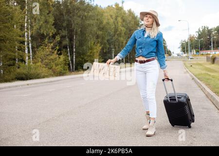 Joyful woman wait passing car on empty road with cardboard poster and suitcase. Lady in hat and denim outfit escape from city by auto stop to go