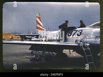One-quarter right side view of a Lockheed F-80C Shooting Star (s/n 49-1873) parked in a revampment, somewhere in Korea. The aircraft bears the markings of the 36th Fighter Bomber Squadron (FBS). One man stands on each wing, removing a top panel to conduct maintenance work. A ladder extends from the right wing to the ground. Another F-80C Shooting Star is parked behind it. Stock Photo