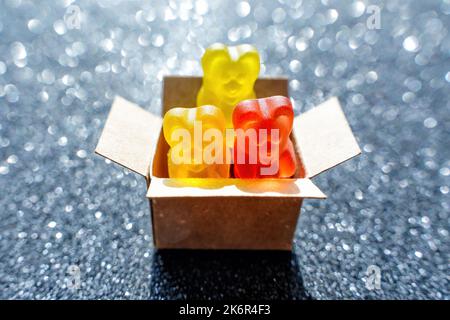 Close-up of three colorful bear shaped gummy candies in a tiny cardboard box against a dark glittering background. Stock Photo