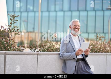 Smiling old business man using digital tablet standing outside city buildings. Stock Photo