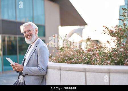 Smiling older business man using digital tablet standing outside city buildings. Stock Photo