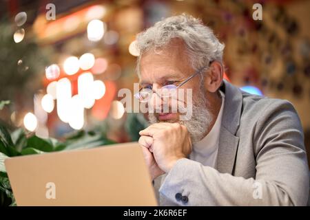 Older business man wearing eyeglasses looking at laptop watching webinar. Stock Photo