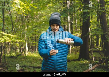 Hiker on a long trail takes a rest and pours hot tea from a thermos into a mug to warm up and replenish vitamins and fluids. High mountain hiking. Stock Photo