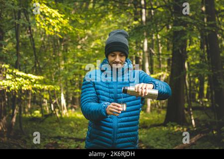 Hiker on a long trail takes a rest and pours hot tea from a thermos into a mug to warm up and replenish vitamins and fluids. High mountain hiking. Stock Photo