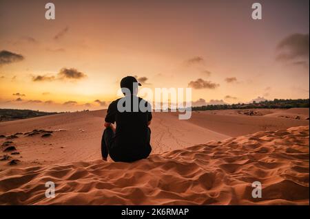 Back view of young asian man wearing hat sitting on sand dune during the sunset at Muine, Vietnam Stock Photo