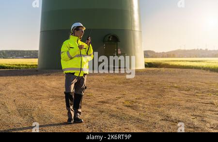 female engineer with safety jacket standing in front of a wind turbine using walkie talkie to checking wind turbine system at a new energy farm Stock Photo