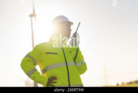 female engineer with safety jacket using walkie talkie to checking wind turbine system at new enegry farm Stock Photo
