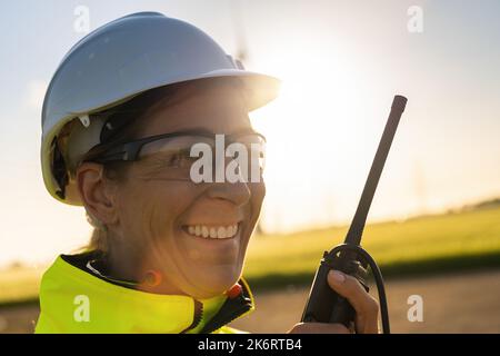 happy female engineer talking in a walkie talkie to checking wind turbine system at a windfarm on sunset Stock Photo