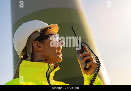 female engineer with safety jacket using walkie talkie to checking wind turbine system at windfarm Stock Photo