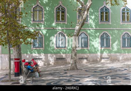 Seated man with tablet on public bench with motor scooter, red  postbox in public square Bario Alto, Lisbon, Portugal Stock Photo