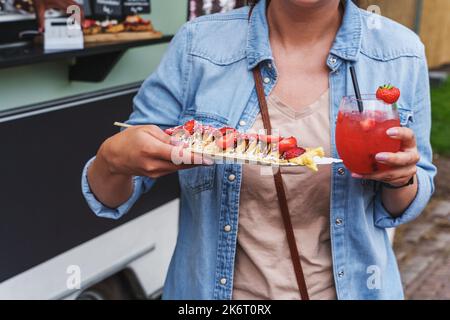 Woman enjoys delicious pancakes with strawberries and strawberry daiquiri at a food festival. Crepes with fresh strawberries and whipped cream, street Stock Photo