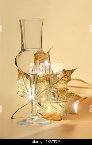 Golden Autumn background. Gilded gold maple leaf distorted by water in small shot grappa glass. Monochrome abstract Fall background, sunlight, long sh Stock Photo