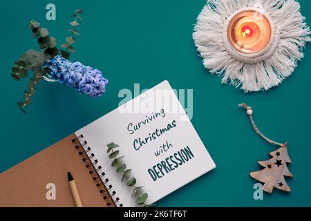 Text Surviving Christmas with depression in notebook, journal. Dark teal, green background, flat lay with eucalyptus, hyacinth flower, Xmas trinket, c Stock Photo