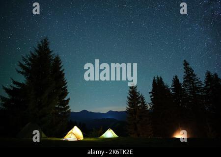 Bright illuminated tourist tents near glowing bonfire on camping site in dark mountain woods under night sky with sparkling stars. Active lifestyle an Stock Photo