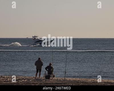 Offshore fishing boat cruising south off the New Jersey coast in the Atlantic Ocean. Fishermen on beach watching the boat with rods in sand. Stock Photo