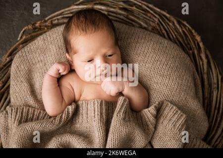 A newborn baby covered with a warm knitted blanket in a wicker basket on a dark background Stock Photo