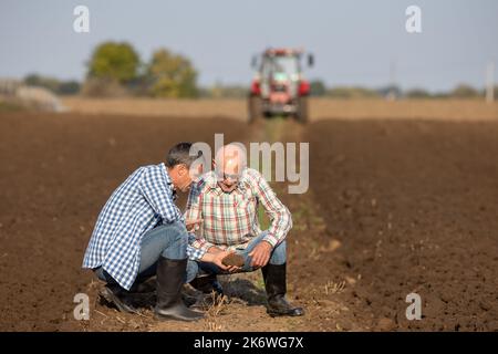 Two mature farmers crouching in field in autumn time, holding clod of earth and checking soil quality while tractor plowing in background Stock Photo