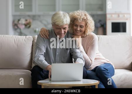Happy older couple sit on couch staring at laptop screen Stock Photo