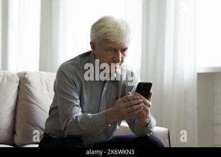 Hoary elderly man spends free time using modern smartphone Stock Photo
