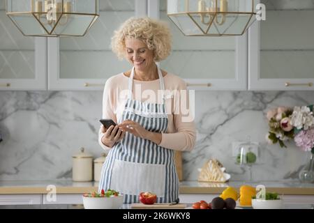Older woman use smartphone standing alone in modern kitchen Stock Photo