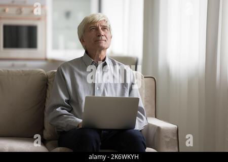 Pensive senior man sits on sofa with laptop Stock Photo
